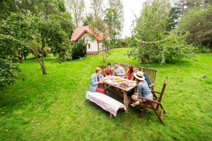 Familj sitter vid dukat träbord i trädgården. I Bakgrundens syns en vit villa.